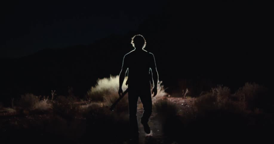 A silhouette of a person holding a guitar stands in a desolate landscape at night with a bright light shining from behind them the light outlines their figure and illuminates some brush and plants on the ground around them