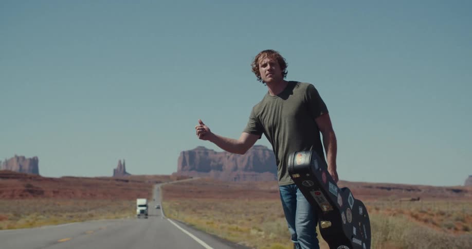A man with curly hair and a green tshirt stands in the middle of a desert road holding a guitar case covered in stickers he looks at the camera and raises his thumb to hitchhike as cars drive by behind him