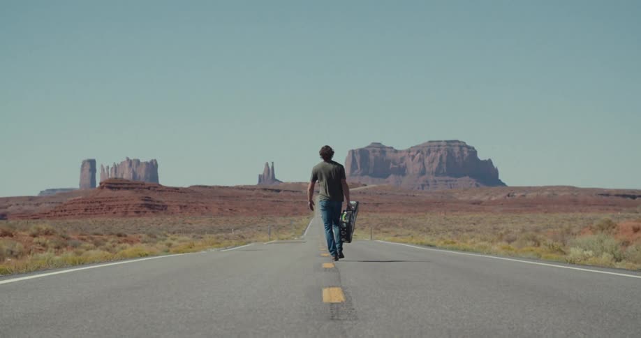 The man carries a guitar case while walking on the road