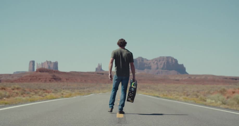 Man walking down a paved street with a guitar in an environment with canyons on the horizon