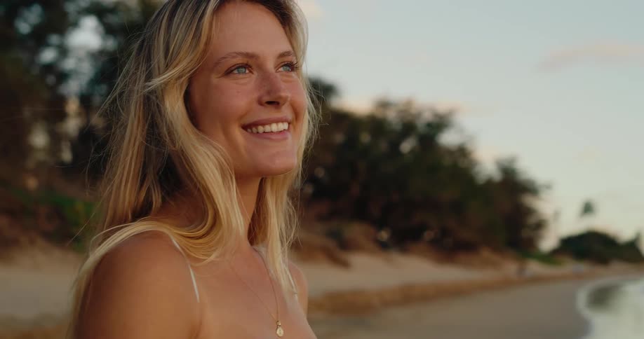 A blonde female smiles warmly while standing on the beach with the shoreline stretching behind her