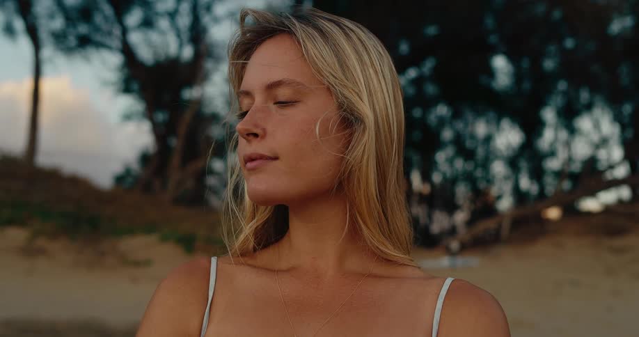 A blonde woman stands on a sandy beach looking out over the water she closes her eyes and takes a deep breath with her shoulders bare and her long hair blowing in the wind