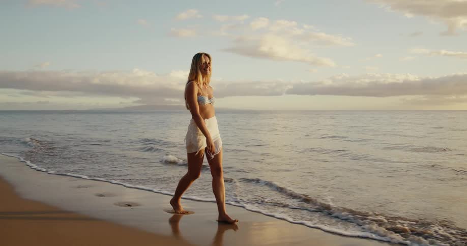 A woman with long blond hair walks and smiles at the camera wearing a light colored bikini top and a white skirt as she passes through the calm water and wet sand on a beach