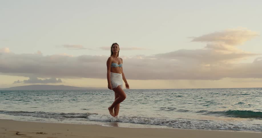 Woman walking on beach along sandy shoreline at sunset
