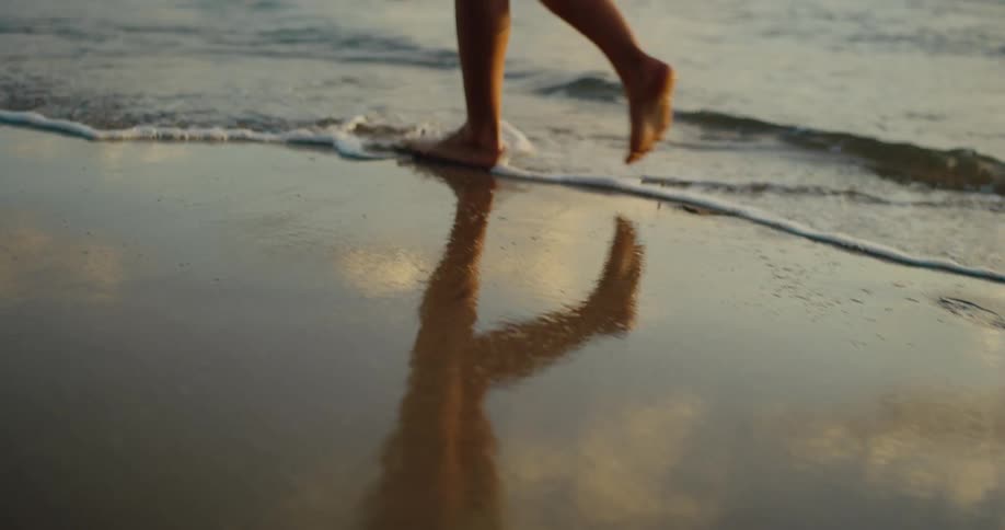 A barefoot person walks along the wet sand of a beach at sunset with their reflection visible in the shallow water as the camera follows their feet the gentle waves of the ocean can be seen lapping at the shore