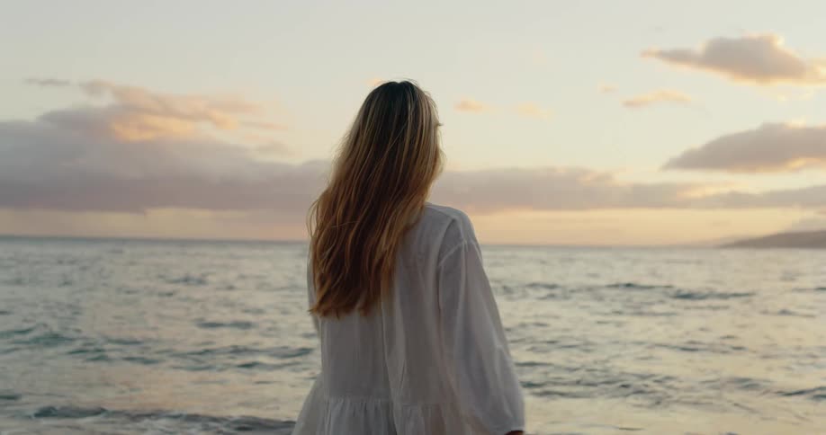 A woman stands on a beach turns around to look at the camera and then turns back toward the water