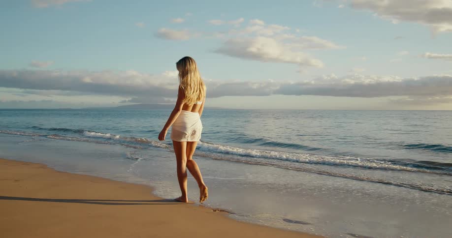 Blonde young woman walking and smiling at the beach next to the ocean outdoors during dusk or dawn