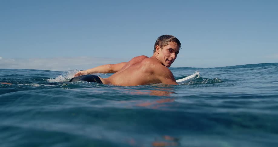 A young male surfer paddles on his board in the open ocean with the camera capturing both above and below the waters surface as he turns to look back