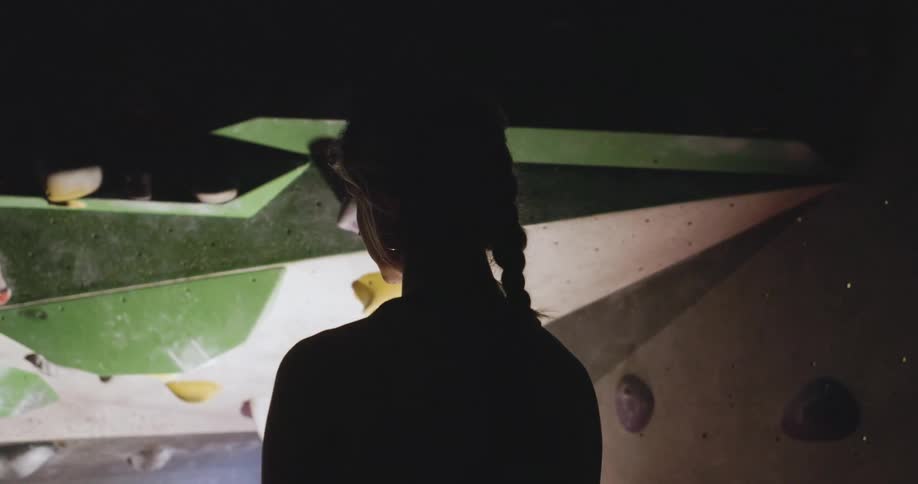 The provided video is only 8 seconds long and shows a static blurry image of the back of a persons head and shoulders the person is standing in a dimly lit space that appears to be a rock climbing gym with green and white climbing walls visible the v