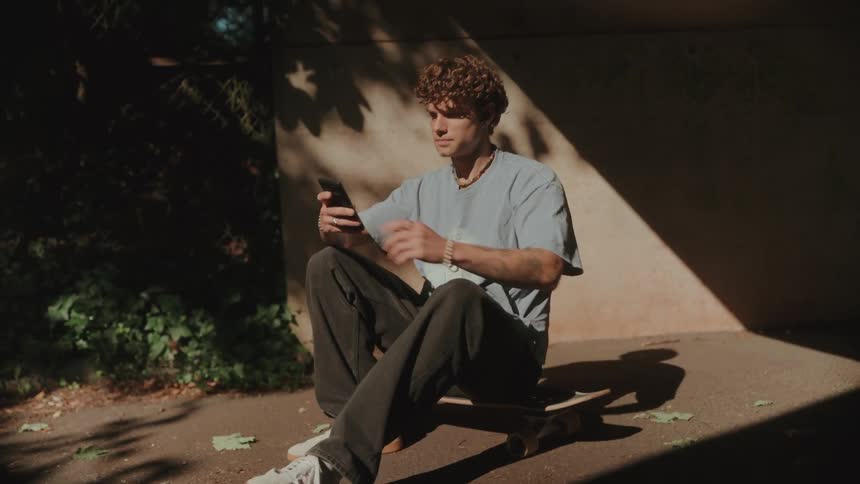 A person sits on a skateboard on the ground and looks at his phone