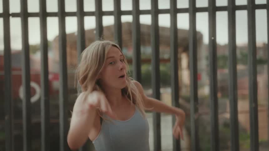 a young blonde woman in a grey tank top dancing in front of a fence