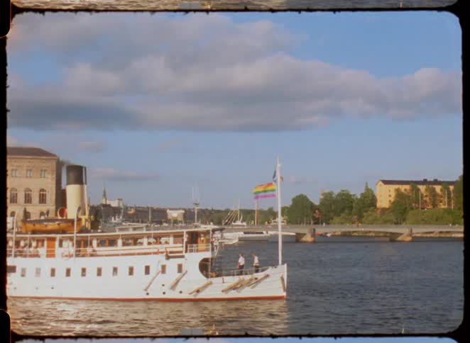 A boat with a smokestack and a flag moves on the water passing a bridge and buildings on the shore