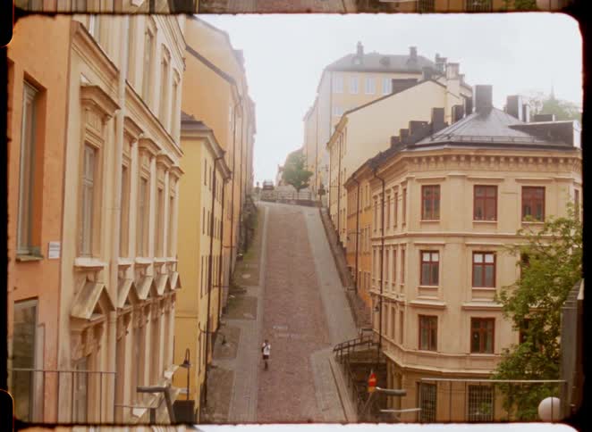 A boy walks down a steep road in a residential neighborhood surrounded by multistory buildings at midday