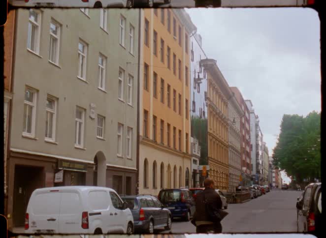 Vehicles parked on a city street lined with tall buildings and a person on a bicycle rides down the street
