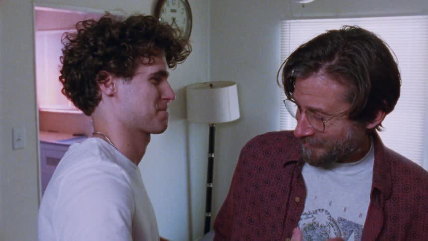A man with a full beard and glasses looks at a teen with curly hair and they both stop talking to hug in a kitchen