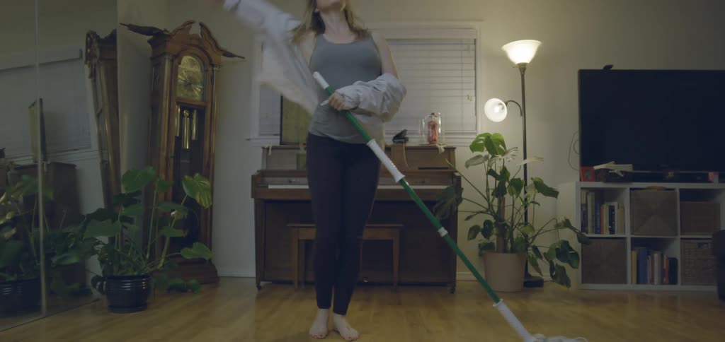 A woman dances while holding a green mop with a white mop head with her head tossed back and her arms in the air in a living room with a piano a grandfather clock and several houseplants
