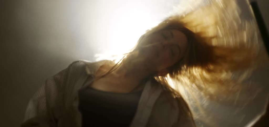 A woman with long blonde hair headbangs in front of a bright glaring light her hair whips back and forth illuminated by the intense light as she moves