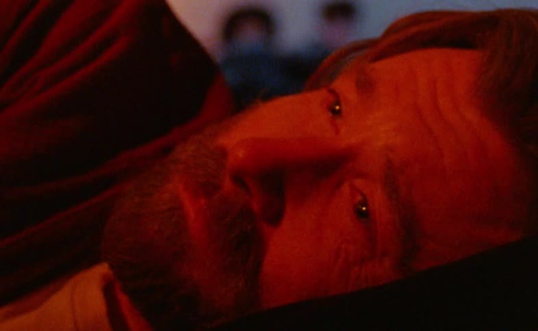 Bearded man lying on his side with a thoughtful expression lit by reddish light