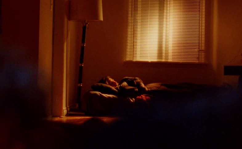 A young woman lies on a couch indoors with a lamp beside her while someone sits opposite her using her phone
