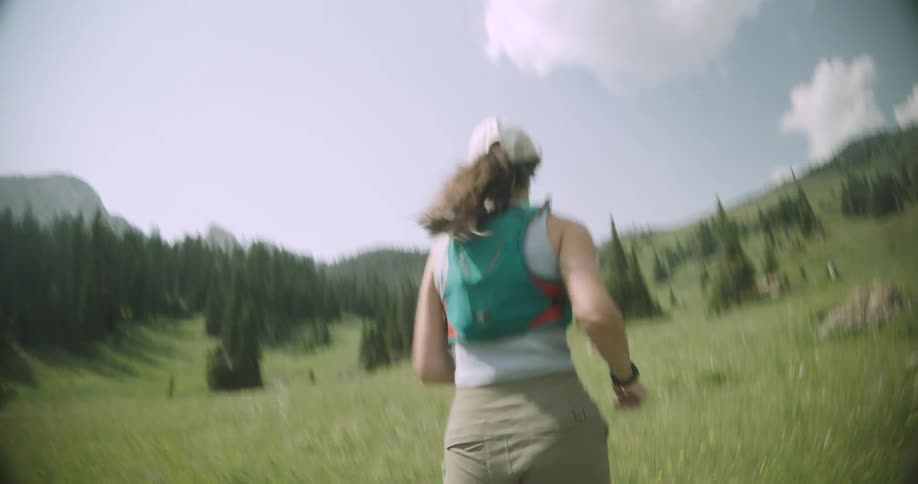 Woman runs through a green field surrounded by mountains and pine trees