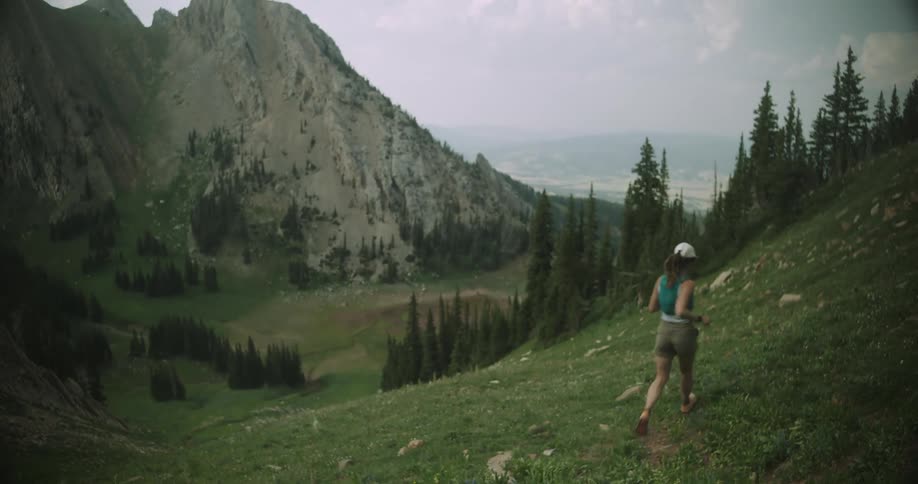 A wide view of a natural landscape shows a person in a white hat a blue tank top and khaki shorts walking down a steep grassy hill in a lush mountainous valley