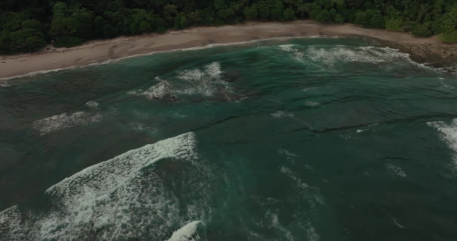 An aerial view of the ocean shows multiple layers of waves crashing towards a sandy beach the coastline is lined with a dense green forest