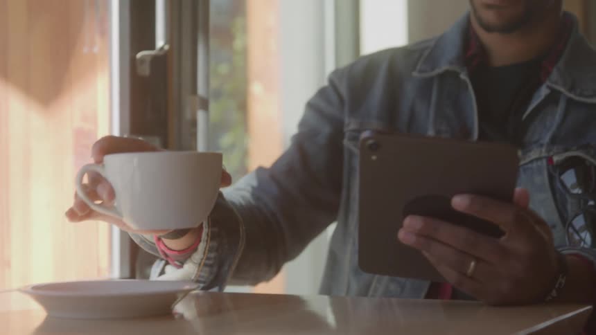 Man in a denim jacket drinks coffee while sitting by the window