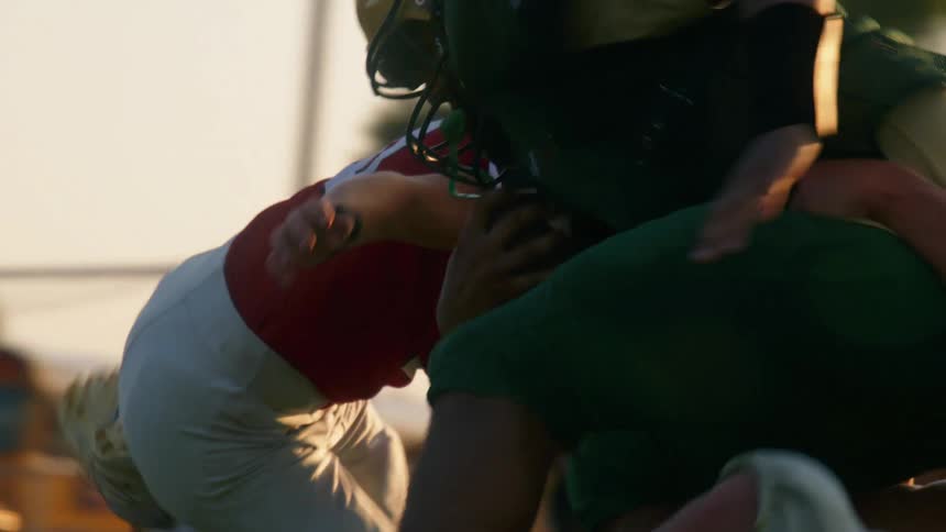 An american football game where a player in a green jersey runs with the ball and is tackled by other players in red jerseys at midday