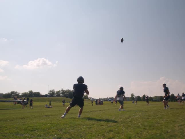 American football players stand on a field in a practice formation on a day with sunlight and clouds