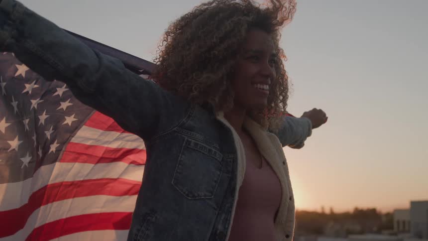A woman with long curly hair stands on a rooftop with her arms spread wide holding a large american flag behind her as a bright sunset illuminates the sky