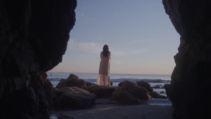 A woman with long dark hair standing on a large rock on a beach as ocean waves crash against the shore