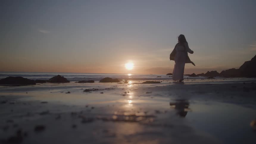 Silhouette of a woman in a white dress spinning on the wet sand of the beach with the sun on the horizon