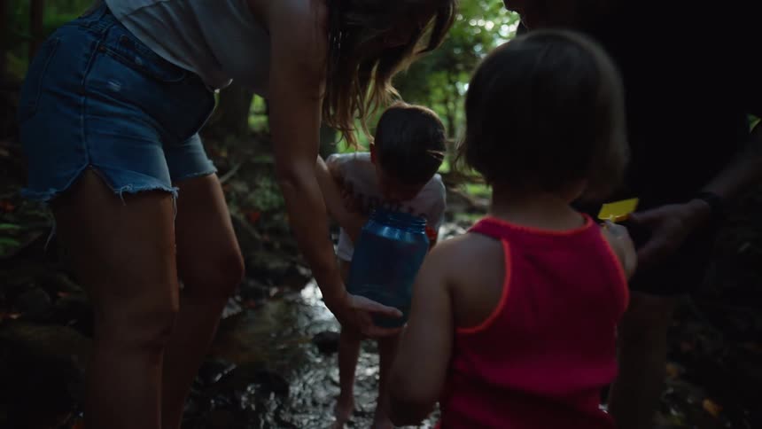 A woman and three children stand and walk barefoot in a shallow stream with the woman and a small boy using a blue water container