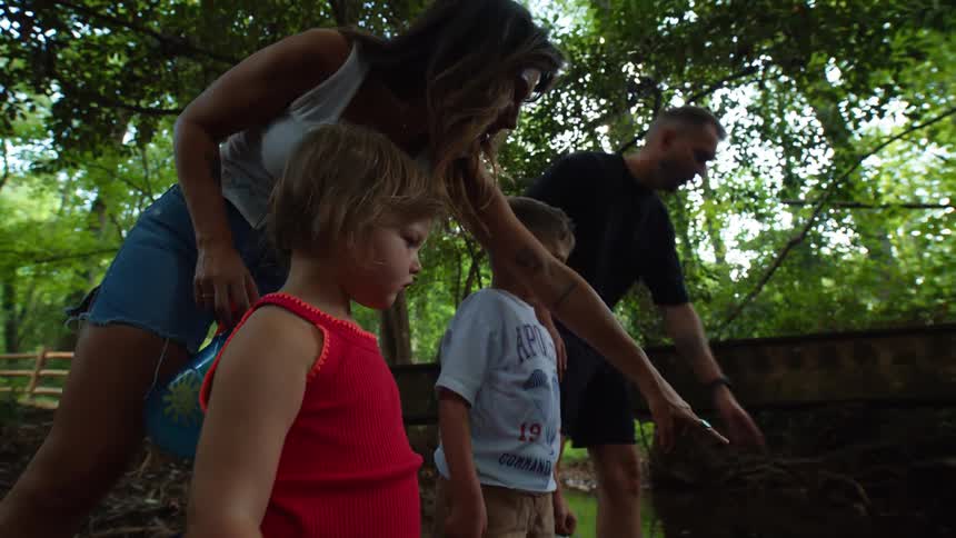 A family stands in a stream with children holding shovels while an adult points at something in the water