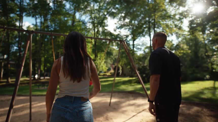Father and mother pushing their children on a swing in a playground surrounded by trees