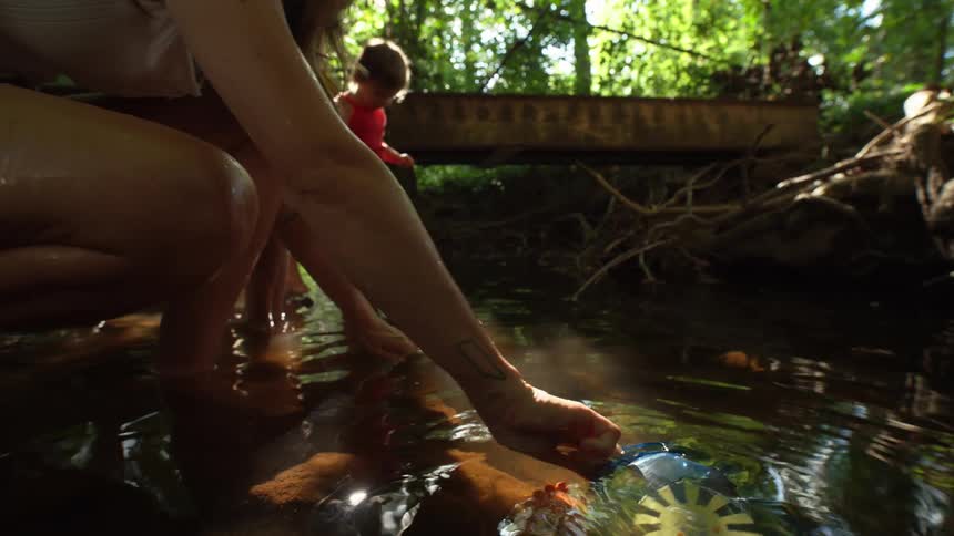 A couple with their young daughter is in the forest as the mother scoops water into a jug from a stream at midday