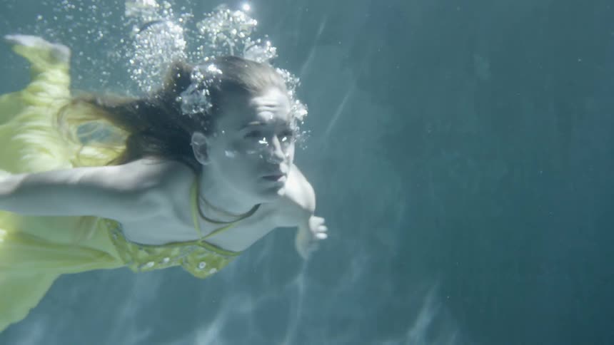 A woman wearing a gown swims underwater in a pool