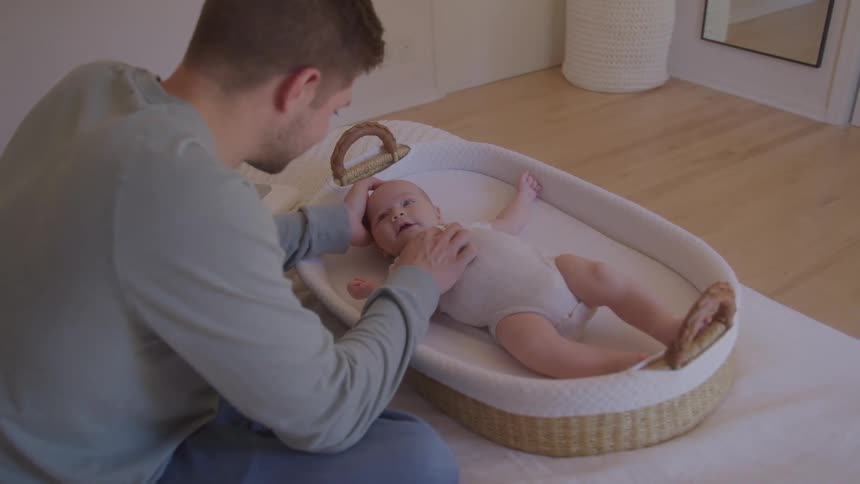 A man wearing a gray shirt and blue jeans sits on a bed and looks down at a baby who is in a wicker bed with white bedding