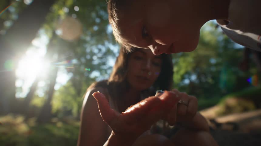Young woman sitting down on a grove next to a forest with her hands covered in mud showing something on her palm to a male child while another person stands by outdoors during day time