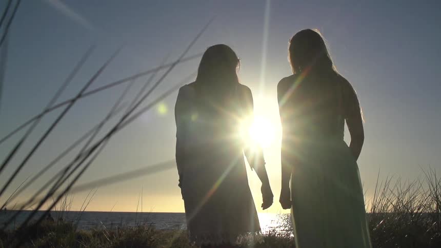 Two women in dresses walking toward the ocean with sunlight shining between them