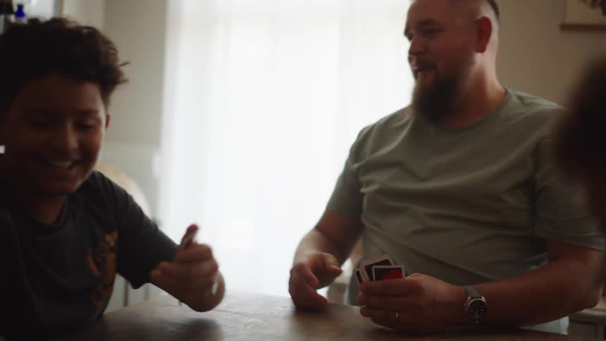 A father with a full beard holds a hand of cards while seated at a table looking down and then smiling as he talks to his family