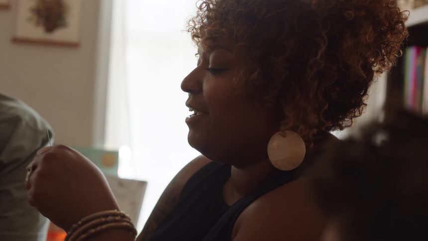 A woman with brown curly hair and large flat earrings holds a card in her hand and laughs as she gestures with the card
