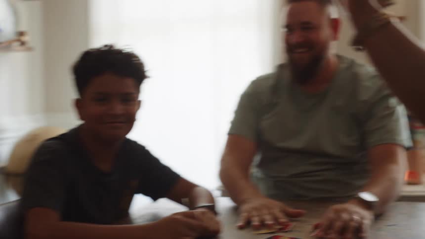 A family of a father mother and three teenage boys sits in the dining room having fun playing a card game as they excitedly highfive each other