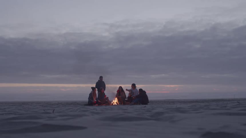 Group of people gathers around a bonfire on the beach at dusk