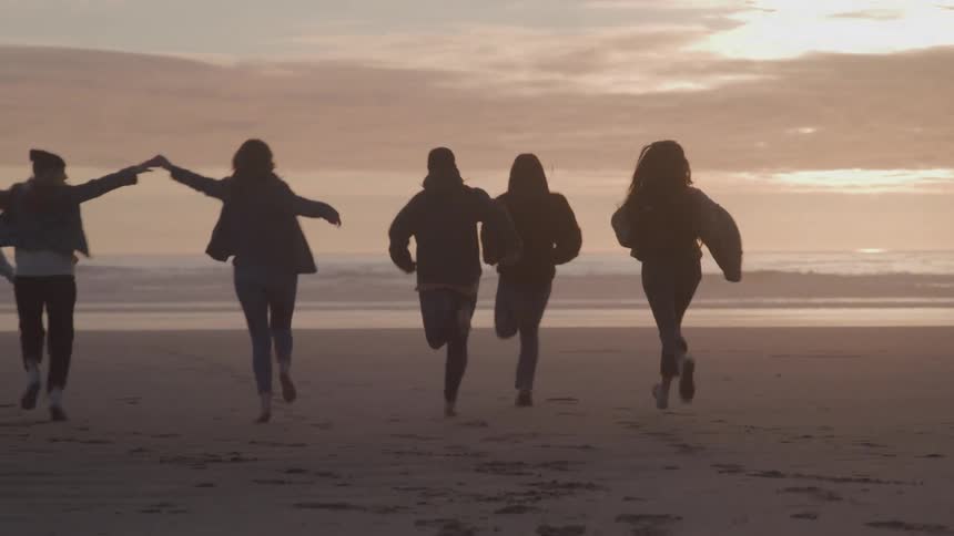 Group of five people runs on the sand towards the sea at sunset