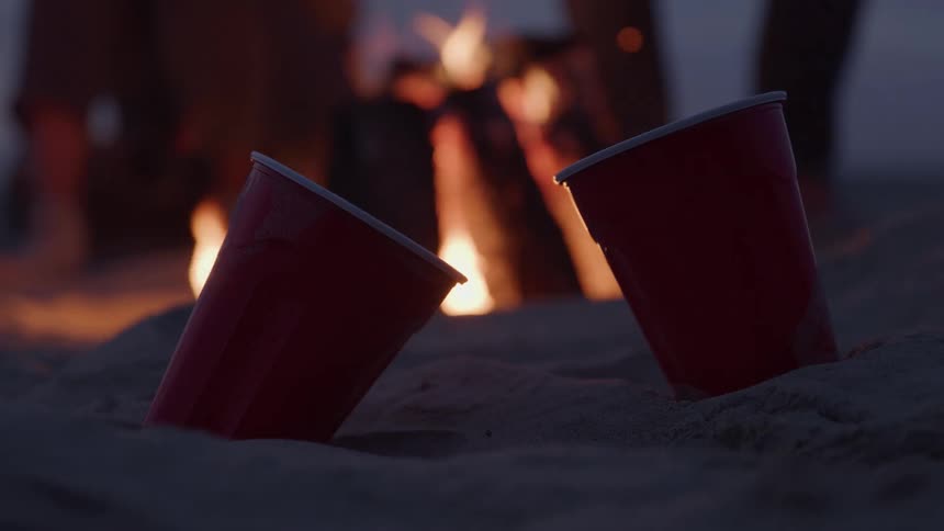 Two disposable drink cups on the sand and in the background a bonfire lit at dusk