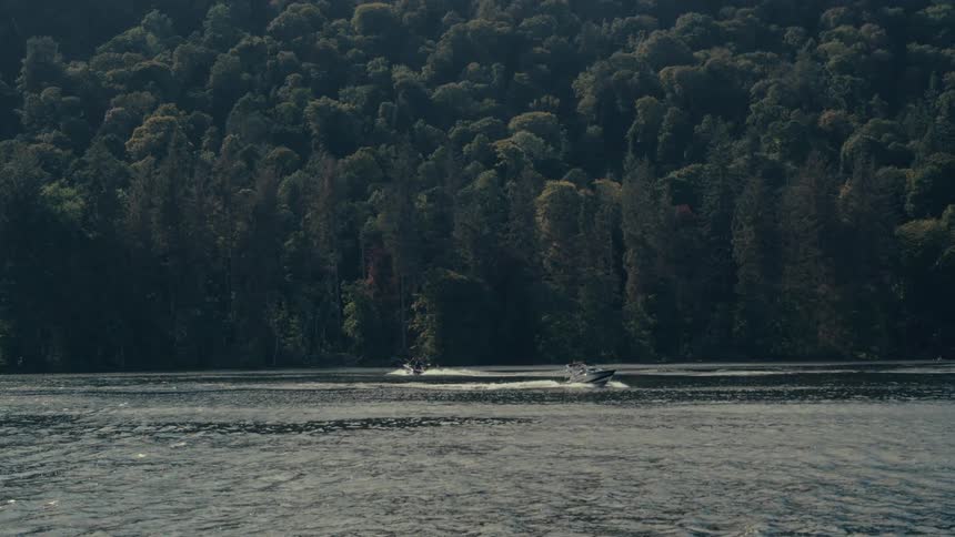 A body of water with a boat sailing from left to right along a forested shore