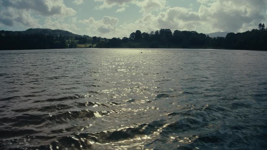 The provided video is a shot of a body of water with sunlight reflecting off the surface in the background there is a shoreline with trees and rolling hills under a cloudy sky