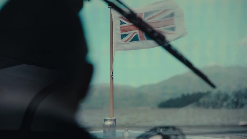 A union jack flag waves from a flagpole on the front of a boat with a persons silhouette in the foreground and distant hills in the background as a windshield wiper moves back and forth