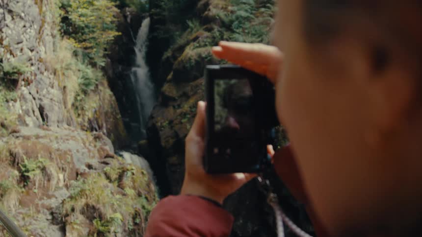 Woman standing in front of a waterfall taking a picture on a digital camera outdoors during day time